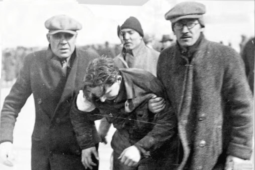 Two men in long coats and caps hold up another man who appears to have difficulty standing