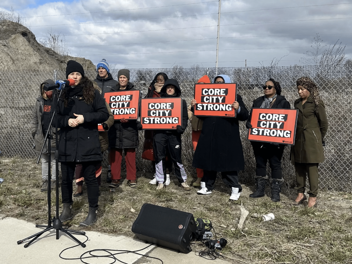 A group of people hold signs reading "Core City Strong" behind a woman wearing black speaking at a mirophone at a press conference regarding the Core City protest.