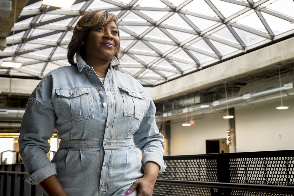A black woman, Deana Neely of Detroit Voltage, smiles and looks into the distance. She's wearing fashionable light denim coveralls in a modern atrium.