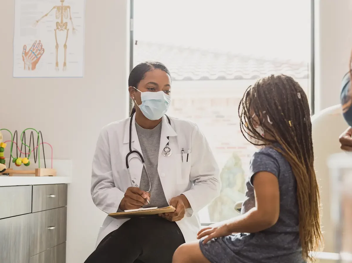 A doctor wears a masks and speaks with a young child in an exam room