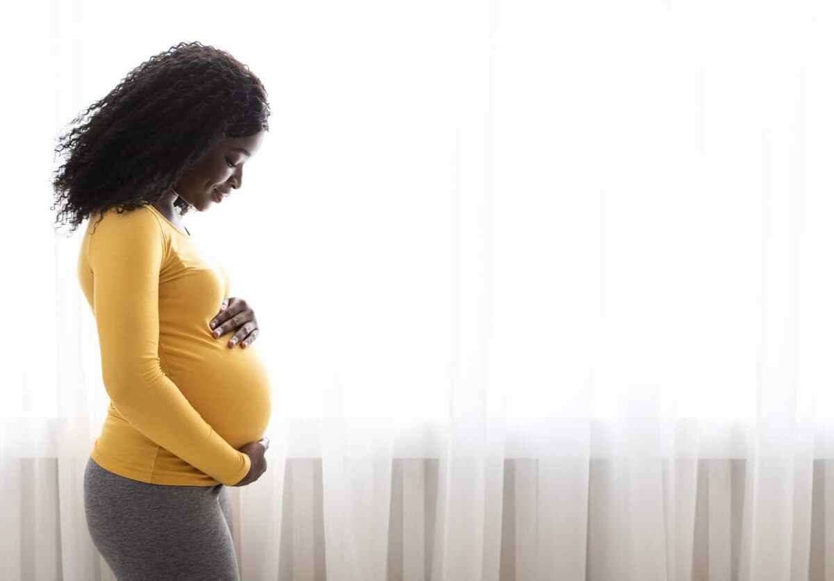 Amid inequities in Black maternal health care, Beautiful pregnant black woman hugging her tummy, enjoying her pregnancy, free space. Side view of african american expecting lady standing next to window at home, touching her big belly.
