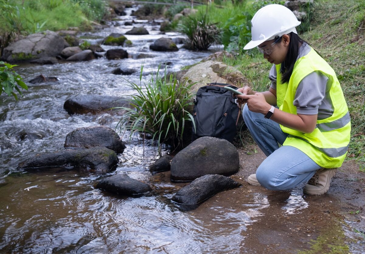 A person in a hard hat and yellow vest kneels beside a stream.