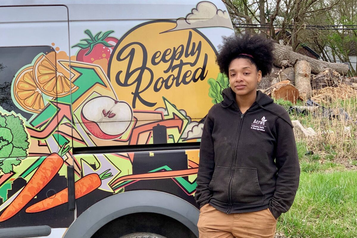 Dazmonique Carr, owner of Deeply Rooted Produce, stands next to a van bearing the company's logo.