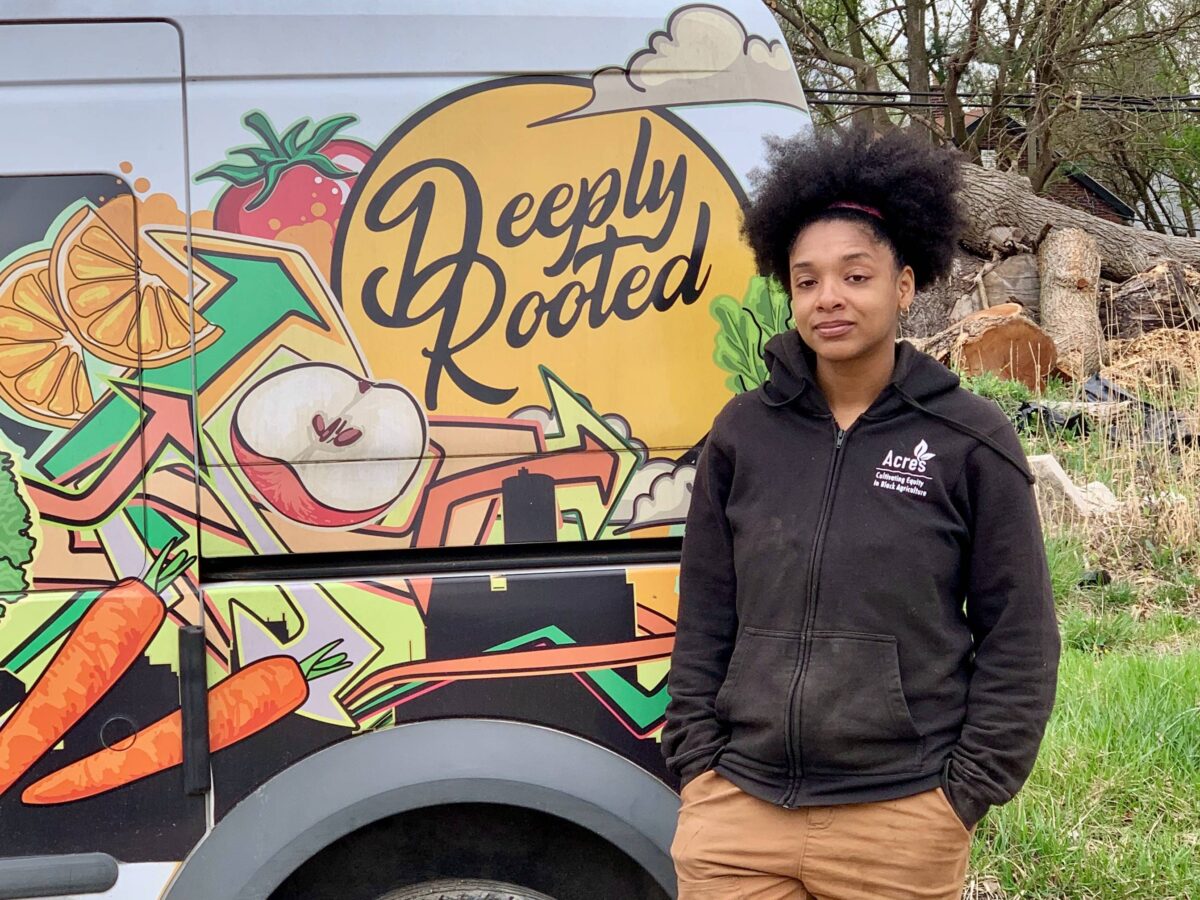 Dazmonique Carr, owner of Deeply Rooted Produce, stands next to a van bearing the company's logo.