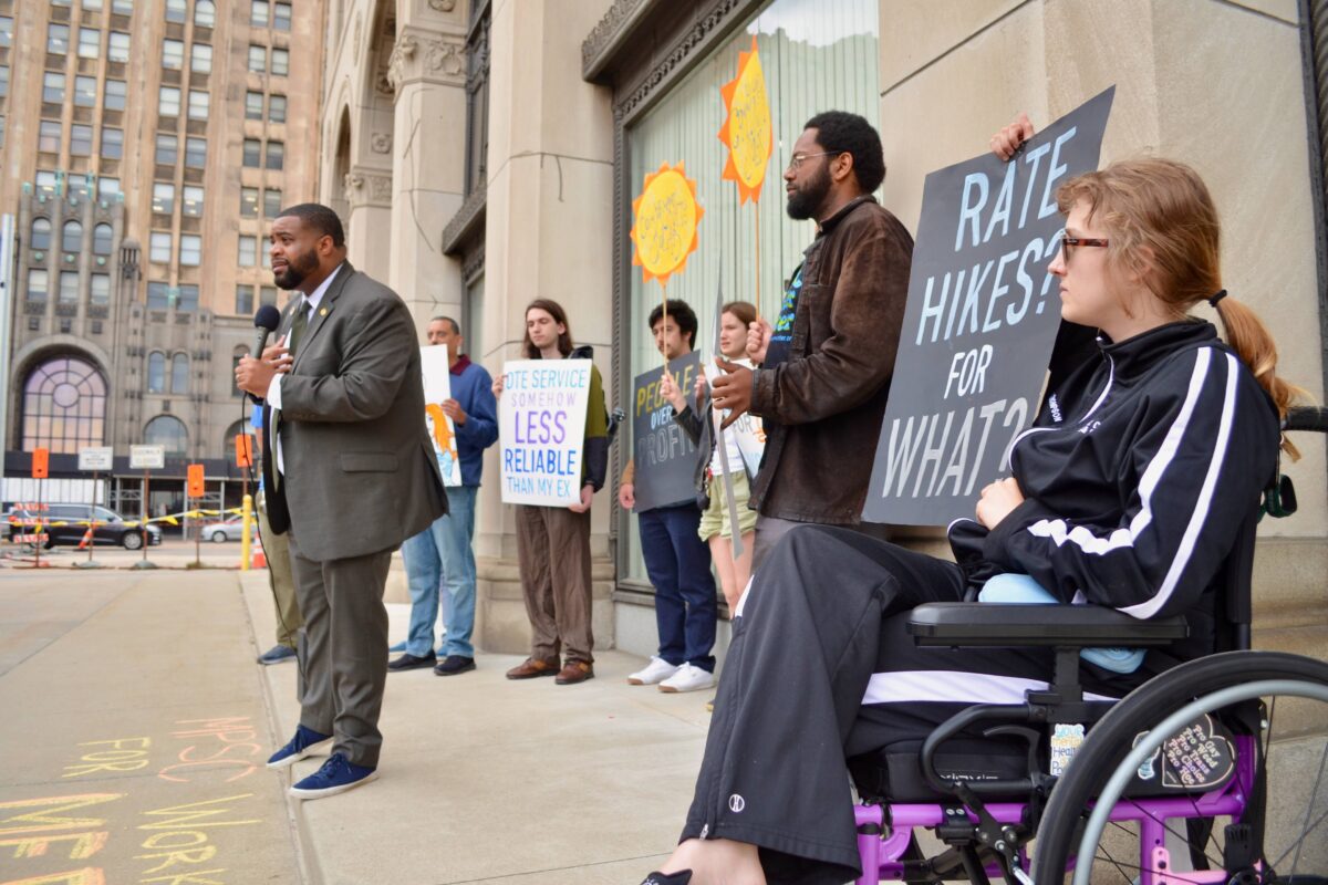 A woman holds a sign stating "rate hikes for what?" as state Rep. Donavan McKinney speaks at a rally outside a Michigan Public Service Commission meeting in Detroit.