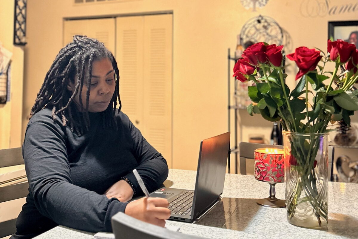 Chicago resident Sherita Hamlin at home, seated in front of a table with a a laptop computer and a vase of roses with a pen in her hand.