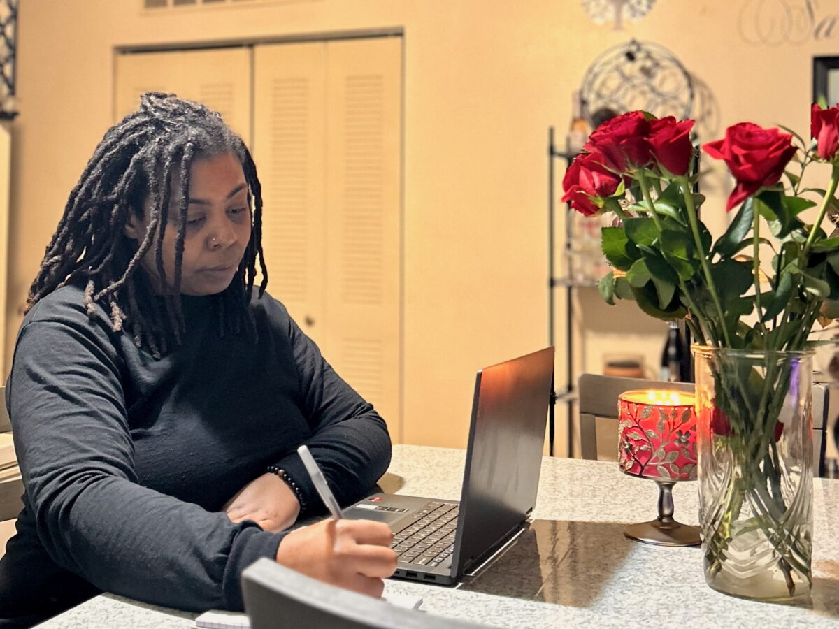 Chicago resident Sherita Hamlin at home, seated in front of a table with a a laptop computer and a vase of roses with a pen in her hand.