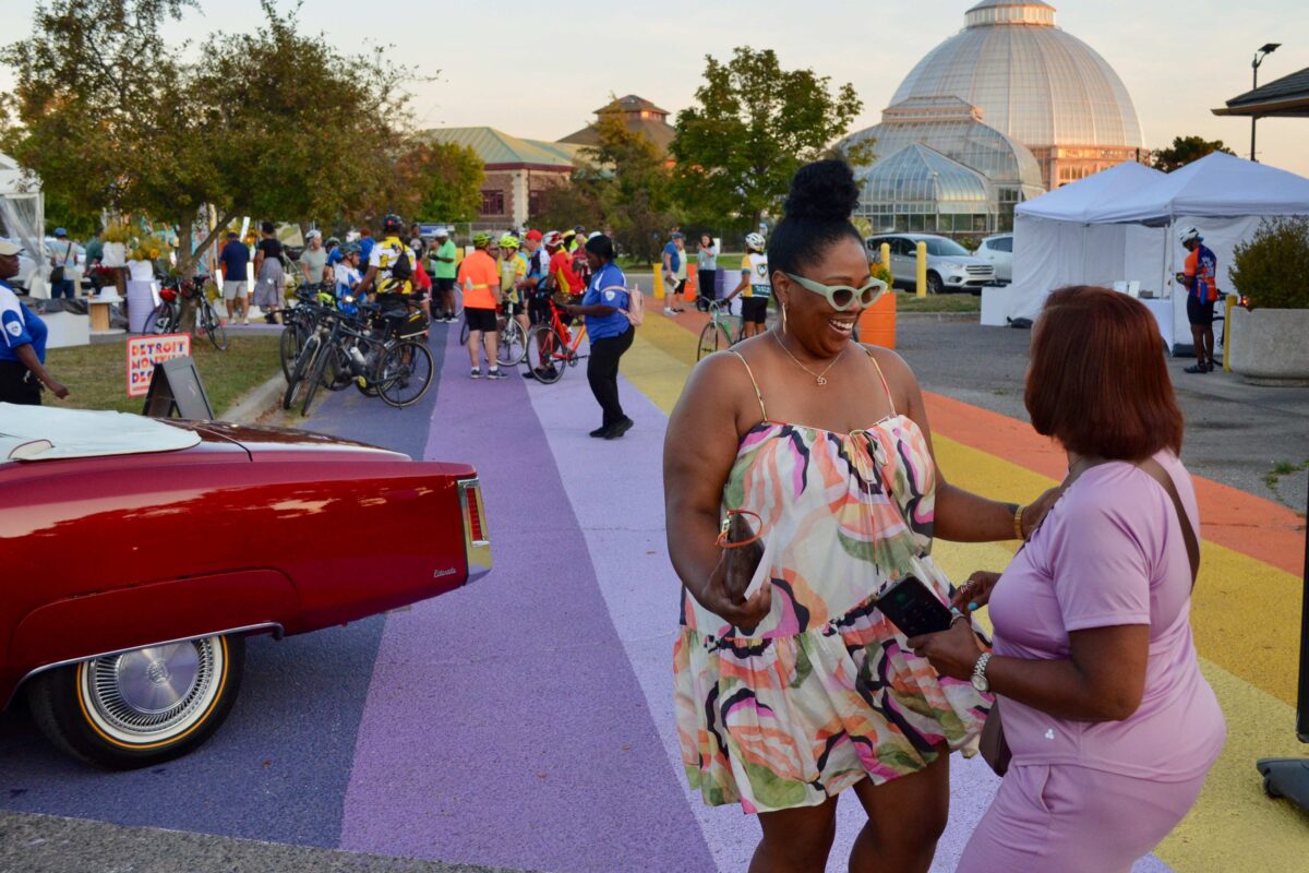 Two women dance near a vintage Cadillac Eldorado, with the dome of the Anna Scripps Whitcomb Conservatory visible in the distance.