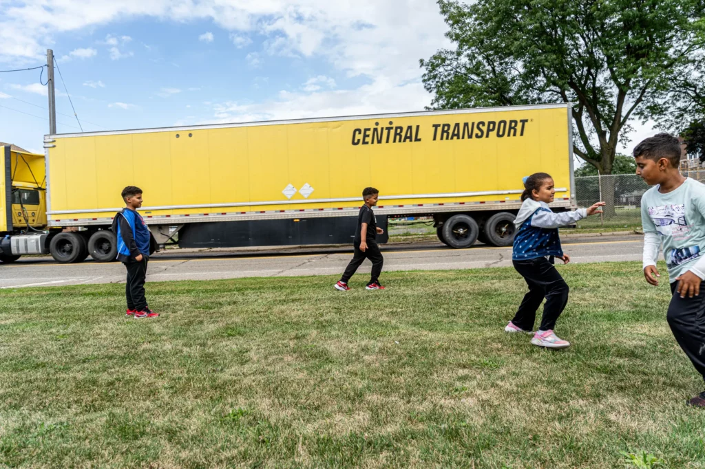 A yellow semi truck passes by on Lonyo Street as children play in the grass.