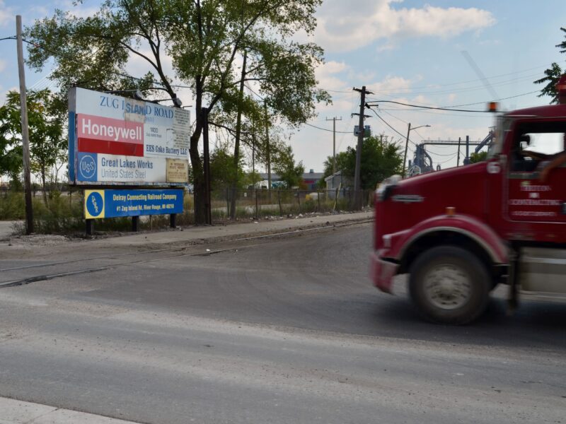 A red truck drives past the Zug Island sign on the corner of West Jefferson Avenue and Zug Island Road.