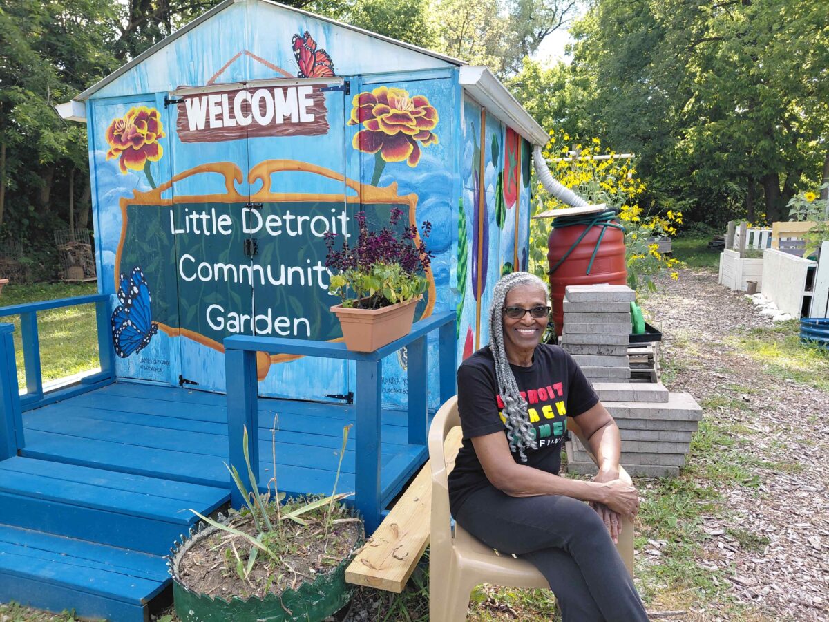 Loretta Powell is seated in front of the painted sign for the Little Detroit Community Garden.