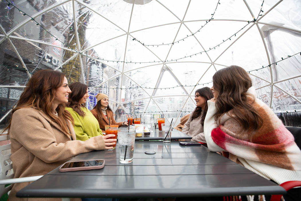 A group of young women sit together in an igloo talking and having drinks.