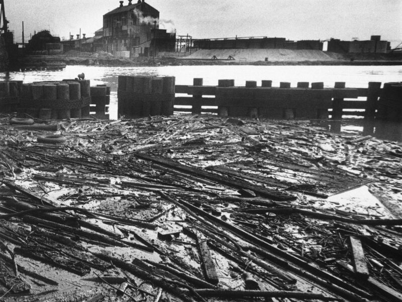 Black-and-white photo of boards, dead fish and other debris lay in an oil slick trapped behind bridge pilings near mouth of Rouge River, May 22, 1969.